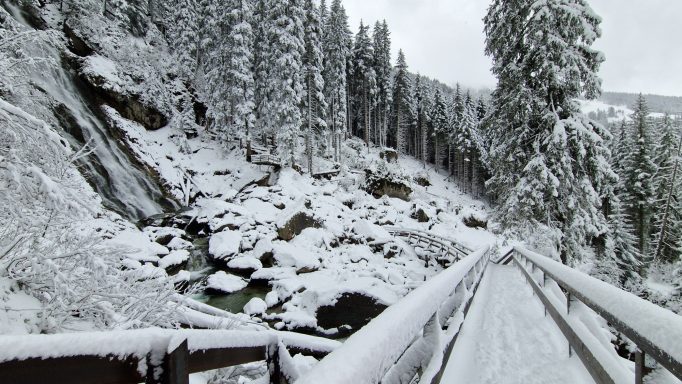 Schneebedeckte Landschaft mit einem Wasserfall und einer Holzbrücke in den Bergen.