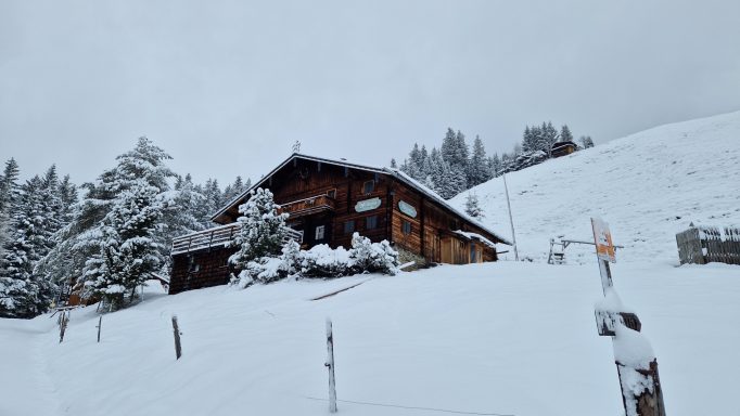 Eine verschneite Hütte in einer winterlichen Landschaft mit Bäumen und sanften Hügeln.